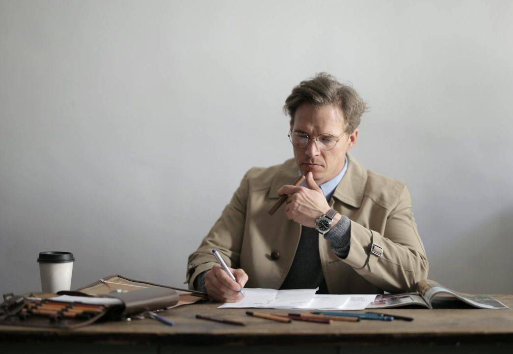 Concentrated male entrepreneur in formal wear eyeglasses and wristwatch holding cigar and writing in papers while drinking takeaway tea in creative studio